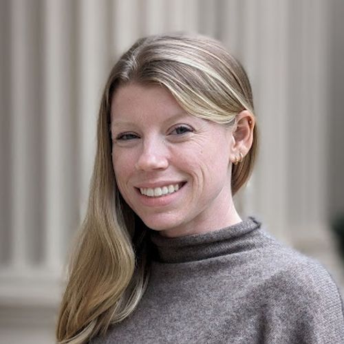 Smiling woman with long blonde hair wearing a gray sweater, standing outdoors in front of large stone columns.